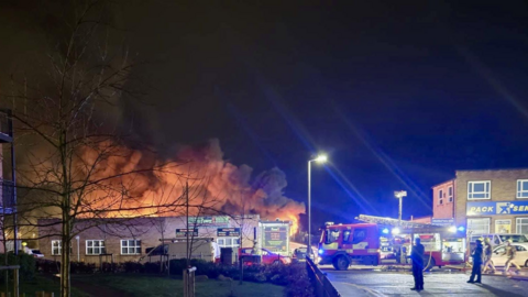 A picture of a well-developed fire in a single-storey industrial building at night. An orange glow comes from the building and it is dark as a fire engine is parked in front of it.