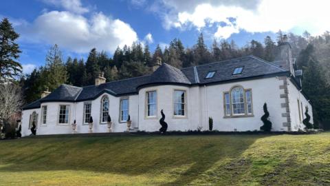 Large white country house with curved bay windows and slate roof, set on a sloping lawn with manicured shrubs, backed by dense woodland under a partly cloudy blue sky.