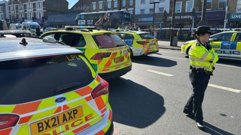 A line of police and ambulance vehicles in a road, with a uniformed police officer standing in front of the vehicles. A row of shops can be seen in the background