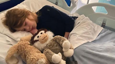 Winifred is pictured asleep in a hospital bed, holding two soft toys under a blanket, with hospital rails visible around the bed.