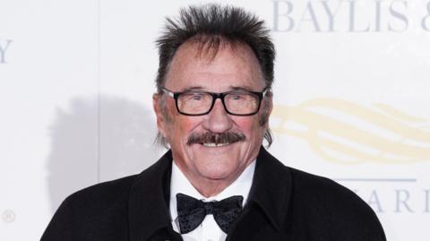 Paul Chuckle at a red carpet event. It is a close crop of his face. He has a brown moustache, brown hair sticking up around his head and black square glasses. He is smiling and wears a white shirt, black bowtie and black jacket.