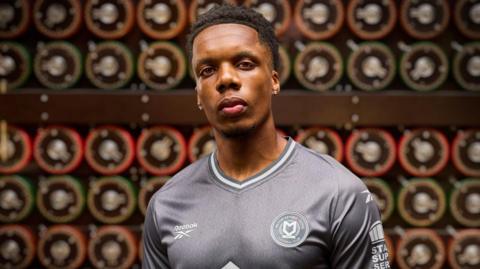 A man is stood in front of a Bombe machine wearing the new grey MK Dons kits