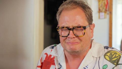 A close up of Alan Carr pulling a pained, scrunched up facial expression.