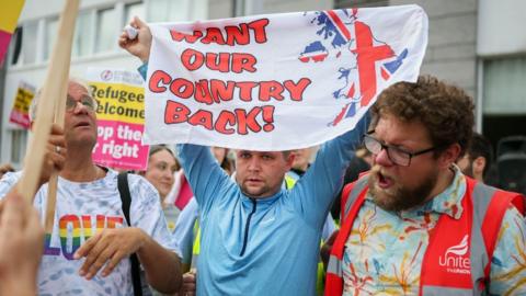 An image of an outdoor protest. A man in the middle, wearing a blue jumper, holds a sign above his head reading "want our country back", with an image of Britain and Northern Ireland coloured in the colours of the union flag. A man to his left is shouting with his mouth wide open, while a man on his right is wearing a t-shirt with "love" in rainbow colours on the front. 