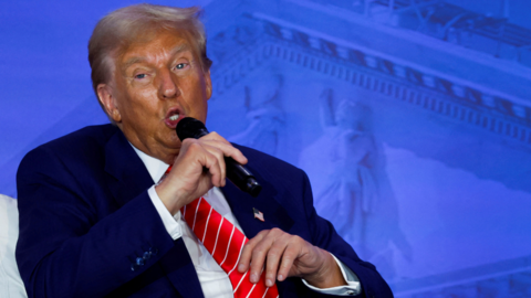 Donald Trump in blue suit, white shirt and red tie talks into a microphone 