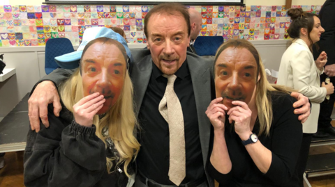 A man with short dark hair and goatee beard, wearing a grey suit jacket and cream tie, is standing between two people holding masks to their face of his face.