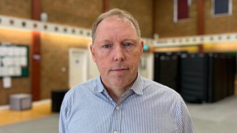 Paul Smith is wearing a blue and white striped shirt and is looking at the camera. He is standing in front of an out-of-focus brick training hall.