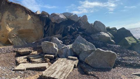 A large mound of rocks on a coastline on a sunny day. There are rectangular bits of broken wall, made of house bricks, in the foreground.