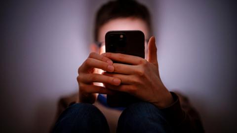 A young person holding a smartphone in a dark room. We cannot see their face but it is illuminated by the phone's screen.