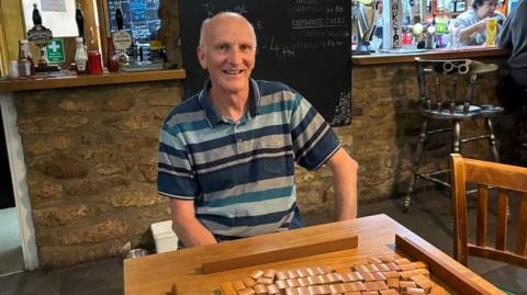 Mark Trenchard set up Shipham's Mahjong club. He is a slim, middle-aged white man who is wearing a blue striped top. He is sitting in a pub with a Mahjong table in front of him. 