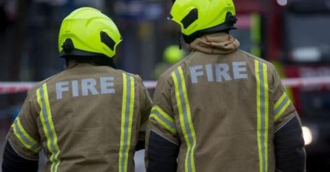 Two firefighters stand with their backs to the camera, wearing yellow fire jackets and helmets. Their heads are tilted towards each other. 