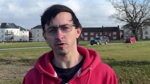 Jordan Crawford ina red hoodie standing on a large area of open grass, with a road and houses and blocks of flats in the distance behind him.
