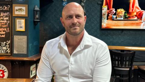 Dave Burns is sitting on a stool in the bar of the pub. He is wearing a white collared shirt. A television screen is in the background