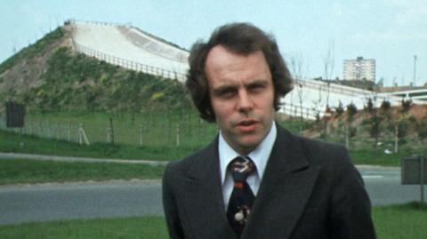 Presenter Martin Young wearing a suit and tie, stood in front of the outdoor artificial ski slope in Kirkby, near Liverpool.