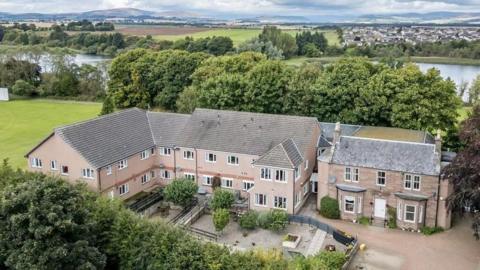A large modern residential building, next to an older stone house. Behind the buildings is a large river and there are hills in the background. 