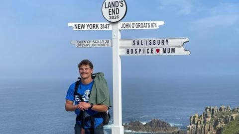 Man stood by a sign at Land's End which shows distances to different locations, with the miles it would take to get there. The sign has New York and John O'Groats on it and also a sign reading "Salisbury Hospice/Mum"