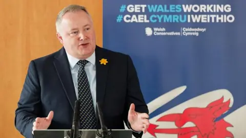 Darren Millar at the Tory party manifesto launch. He has a dark blue jacket over a light blue shirt and a patterned dark blue tie. He has short grey hair and a yellow daffodil on the left lapel of his jacket.