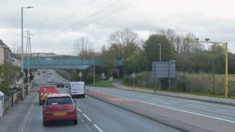General view of a road. Cars are travelling along the road. Trees and houses line either side of it. Power lines are visible above the road.