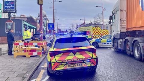 A police car with it's blue lights on parked on a cordoned off road. There are two amublances and an another police car parked beside blue and white police tape.