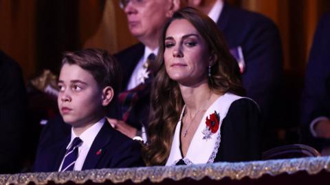 Prince George and the Princess of Wales sitting overlooking the auditorium. They are not smiling and are both wearing poppies