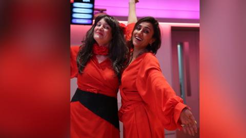 Two women embrace and pose for the camera. They are wearing red dresses like Kate Bush.