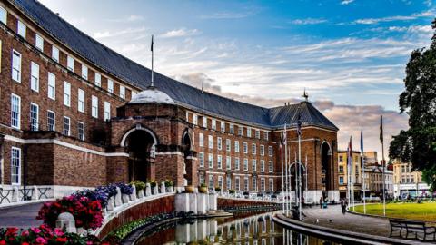 Exterior of Bristol's City Hall. The building has brown bricks and a white bottom. It has water around the perimeter and a green lawn with flag roles.