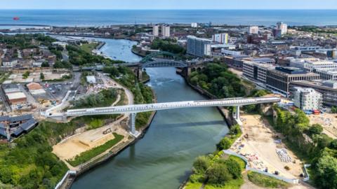 An aerial view of the footbridge. It is a long white structure crossing the River Wear and connecting Keel Square on the right to the Stadium of Light, with cranes next to it on both shores. Wearmouth Bridge and the North Sea are also visible in the distance.
