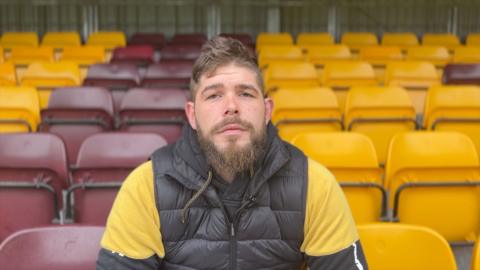 A man with light brown hair and facial hair sits in front of empty stadium seats. He is wearing a black sleeveless jacket and yellow top underneath.