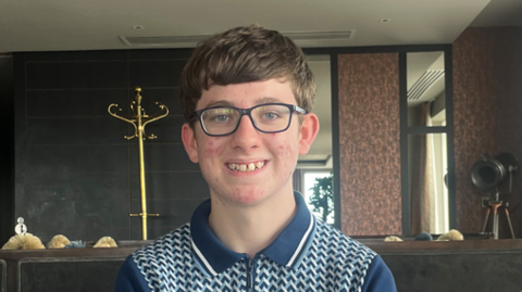 Teenage boy with brown hair smiling in into the camera. He is wearing dark rimmed glasses and a blue patterned top.