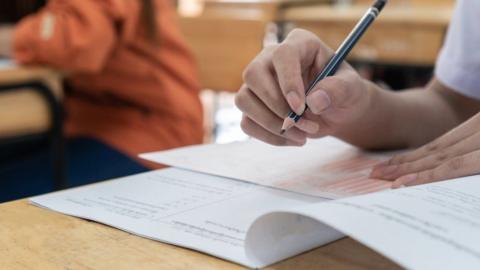 A close-up of a girl's hand is seen writing with a pencil on a test paper in what looks like an exam hall in a school