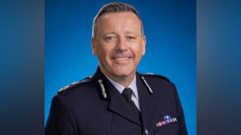 A close-up photo of the acting chief constable of West Midlands Police. He wears a black police uniform and stands in front of a blue background, looking into the camera.

