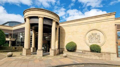 The court has stone pillars at its entrance and two bushes along one wall. The wall has a royal crest on it.