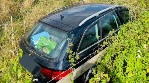 A dark-coloured car shown half-buried in bushes next to the M4. The boot of the car is full of bags and a fence and barbed wire is visible in the foreground. 