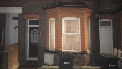 The exterior of the ground floor of a redbrick terraced house, which has a bay window and the curtains drawn, with the light on inside. The front door is to the left and outside are two wheelie bins, with the word "Luton" on the front.