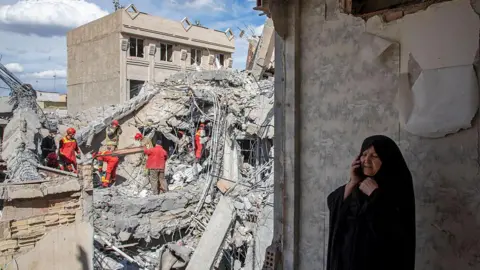 A woman speaks on the phone as emergency workers sift through rubble in Tehran