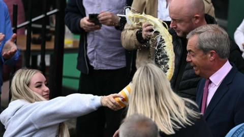 A woman in a grey hoody is holding a paper cup in her outstretched hand. There is yellow milkshake coming out of it, flying through the air in the direction of Nigel Farage. He is wearing a dark suit with pink shirt and tie. 