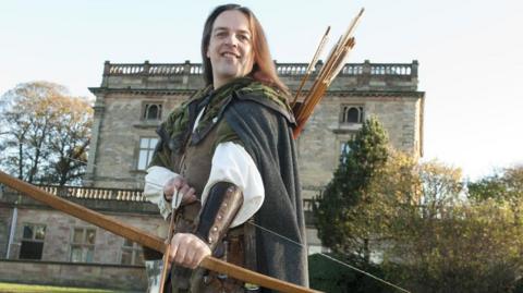 A man with long hair and green robes holding a bow and arrow. He stands in front of Nottingham Castle