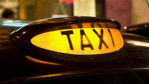 A close-up shot of a lit-up yellow taxi sign on the top of a car. It says "TAXI" in bold across it. The background is blurred.
