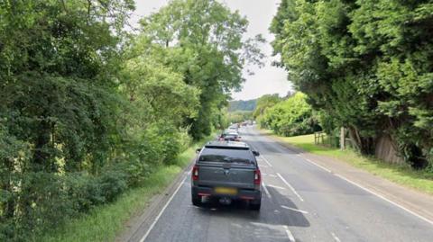 A main road goes through countryside with trees and fields on both sides. There is a pavement on one side of the road. The Google Maps image shows a line of cars travelling one way and the road is clear on the other side.