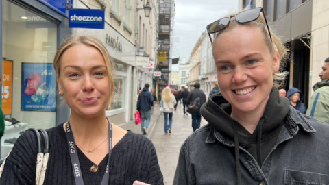 Two young women standing side‑by‑side on a pedestrian shopping street. The person on the left is dressed in a dark ribbed top and wears a lanyard labelled “STAFF". The person on the right wears dark clothing with a hood and sunglasses positioned on top of their head.