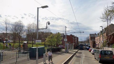 A Google Streetview image of Shipstone Street tram stop in New Basford in Nottinghamshire. The image shows a tram approaching the stop with cars parked on the right of the tram. There is a children's play area on the left of the the image, behind the tram stop. 
