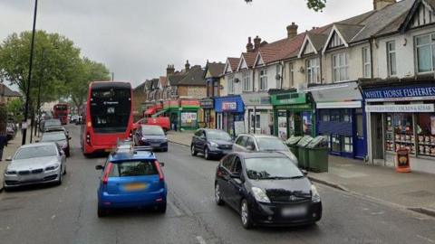 A black car is driving facing forward while a blue car and red bus are driving away from High Street South in East Ham. Cars are parked alongside and there is a row of shops to along the right side of the picture and trees on the left.