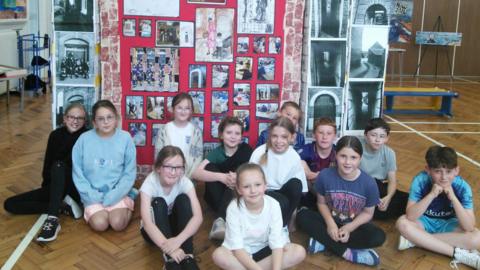 Twelve primary school children sitting on a wooden herringbone floor in front of large display of artwork.