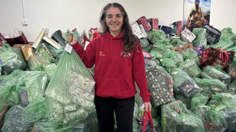 Jo Barrow has long brown hair and is wearing a red hoodie. She is holding a green bag full off wrapped Christmas presents. She is standing in front of a pile of hundreds more green bags that are also filled with Christmas presents.