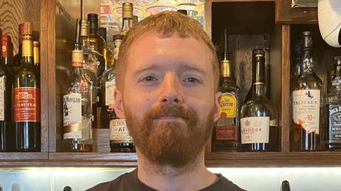 Rhys Vivian has short ginger hair and a ginger beard. He is standing at the back of the bar, with bottles of spirits and wine behind him.