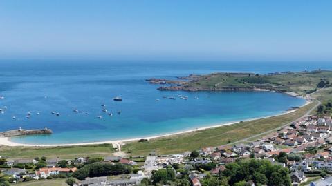 A view of Newton in Alderney - the c curve of the island looking out onto the ocean with fishing boats docked in the sea. Blue skies.