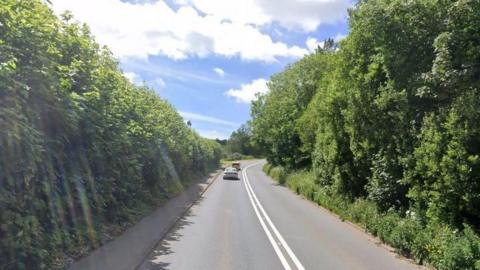 Two cars - one coloured white and the other yellow - drive along the tree-lined A381 near Totnes in Devon on a sunny day.