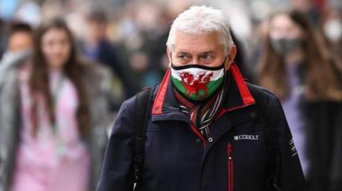 Man stood in crowded street wearing a mask with a Welsh flag on it. He is wearing a blue coat and has a rucksack on. People can be seen out of focus behind him.