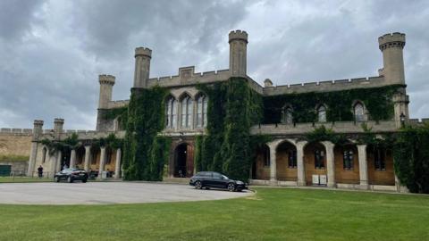 Outside of Lincoln Crown Court, a two-storey stone building in Gothic style with crenelated turrets and vegetation growing around the wooden doors.