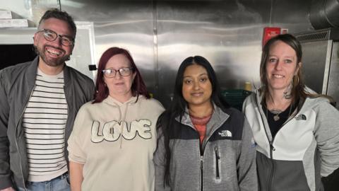 A row of four people looking at the camera - on the left is a man with curly grey hair glasses, a stripy top who is smiling with his teeth. Next is a woman with red hair and glasses wearing a beige jumper with the word 'LOVE.' Next is a woman with dark hair and a North Face jacket on, and on the right is a woman with neck tattoos wearing hoop earrings and smiling with her hands behind her back.